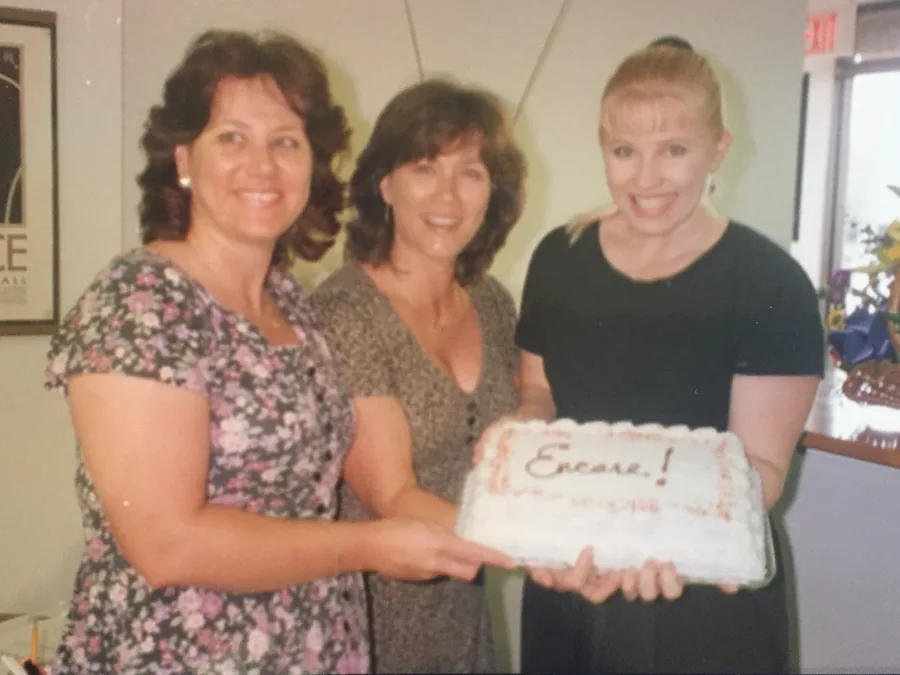 Three women smiling over a sheet cake iced with the word Encore