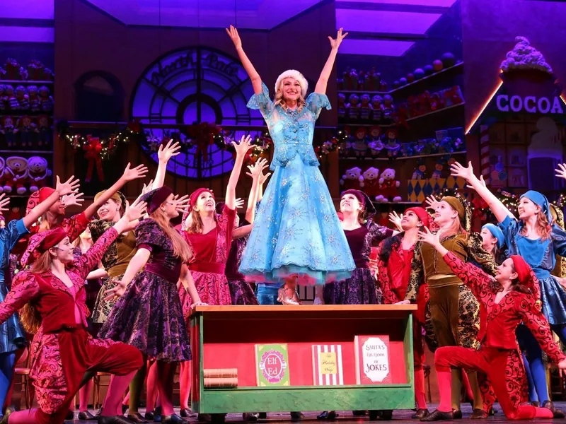 A performer in a blue gown stands on a desk in Santa's workshop, surrounded by elves reaching toward her